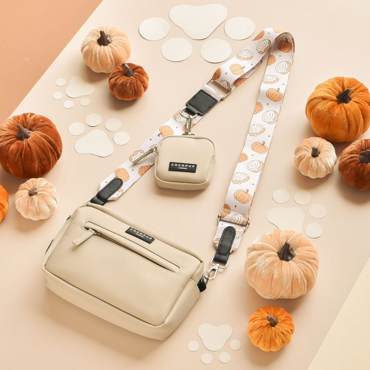 A beige crossbody bag and matching small pouch are displayed on a two-toned peach and beige background. The bag strap, named "Cinnamon Buns" by Cocopup London, is perfect for adding a touch of festivity. Surrounding the bags are small to medium-sized pumpkins and white paw print cutouts.