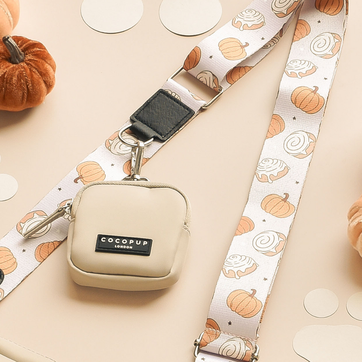 A beige crossbody bag with a Cinnamon Buns patterned strap from Cocopup London lies on a beige surface surrounded by small pumpkins and circular dot decorations. The bag has a metal clasp and features a branded patch reading "Cocopup London." Perfect for adding a festive touch to your Halloween outfit!