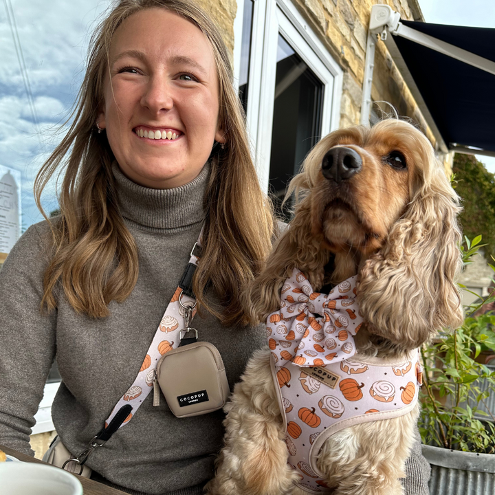 A woman with long hair, wearing a gray turtleneck and adjusting her Cocopup London Bag Strap - Cinnamon Buns, sits beside a brown Cocker Spaniel dog dressed in a pumpkin-themed harness and bow tie. They are sitting outside near a window, with greenery and part of the building visible behind them. Both are smiling.