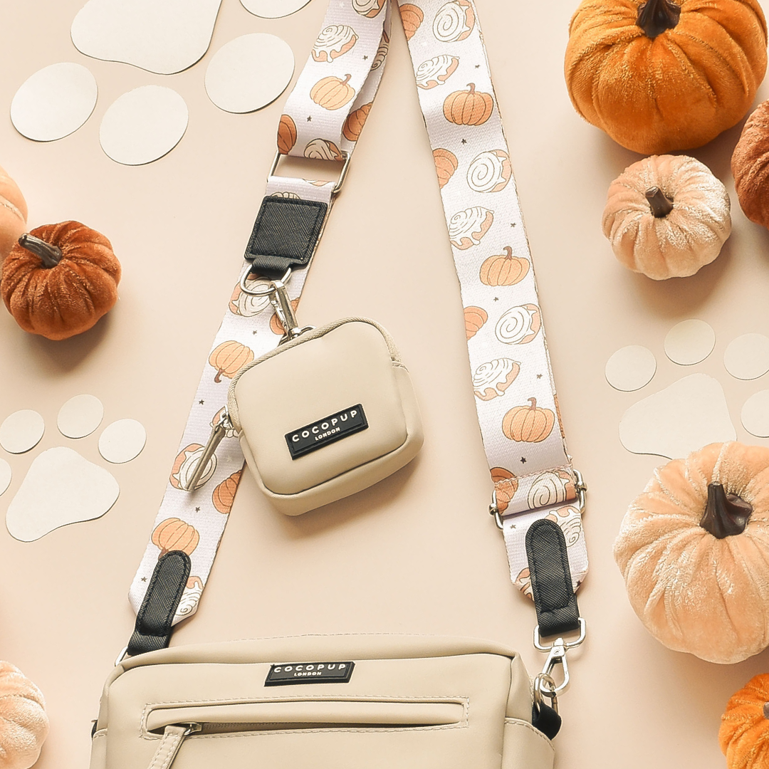 A beige crossbody bag with the Cocopup London Bag Strap - Cinnamon Buns, featuring a pumpkin-patterned design and an attached small pouch, lies on a flat surface surrounded by small pumpkins, beige paper circles, and paw print cutouts. The items are arranged in a Halloween-themed setup that feels as cozy as cinnamon buns on an autumn day.