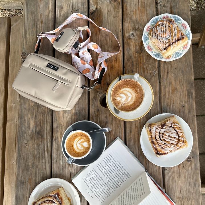 A wooden table holds three lattes featuring intricate latte art, paired with three delicious-looking cinnamon buns on plates. An open book rests beside a beige crossbody bag adorned with the Cocopup London Bag Strap - Cinnamon Buns draped over the chair, enhancing the cozy ambiance. The setting appears to be an outdoor cafe, ideal for a relaxing afternoon.