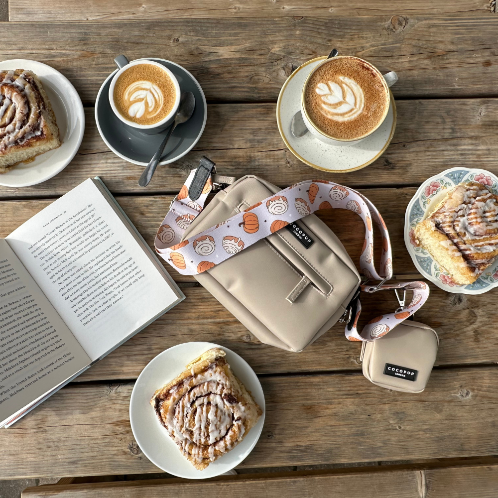 Four plates with cinnamon rolls, three cups of latte art coffee, an open book, and a beige crossbody bag adorned with the Cocopup London "Bag Strap - Cinnamon Buns" on a wooden table. There is also a small beige pouch next to the bag. The scene gives off cozy Halloween vibes, with the cinnamon buns enhancing the festive atmosphere.