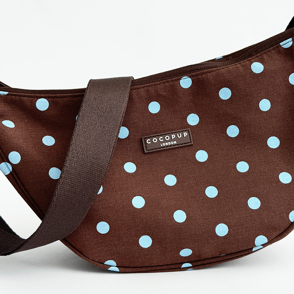 A brown fabric crossbody bag with a light blue polka dot pattern. Featuring Cocopup London logo badge and plain brown strap.