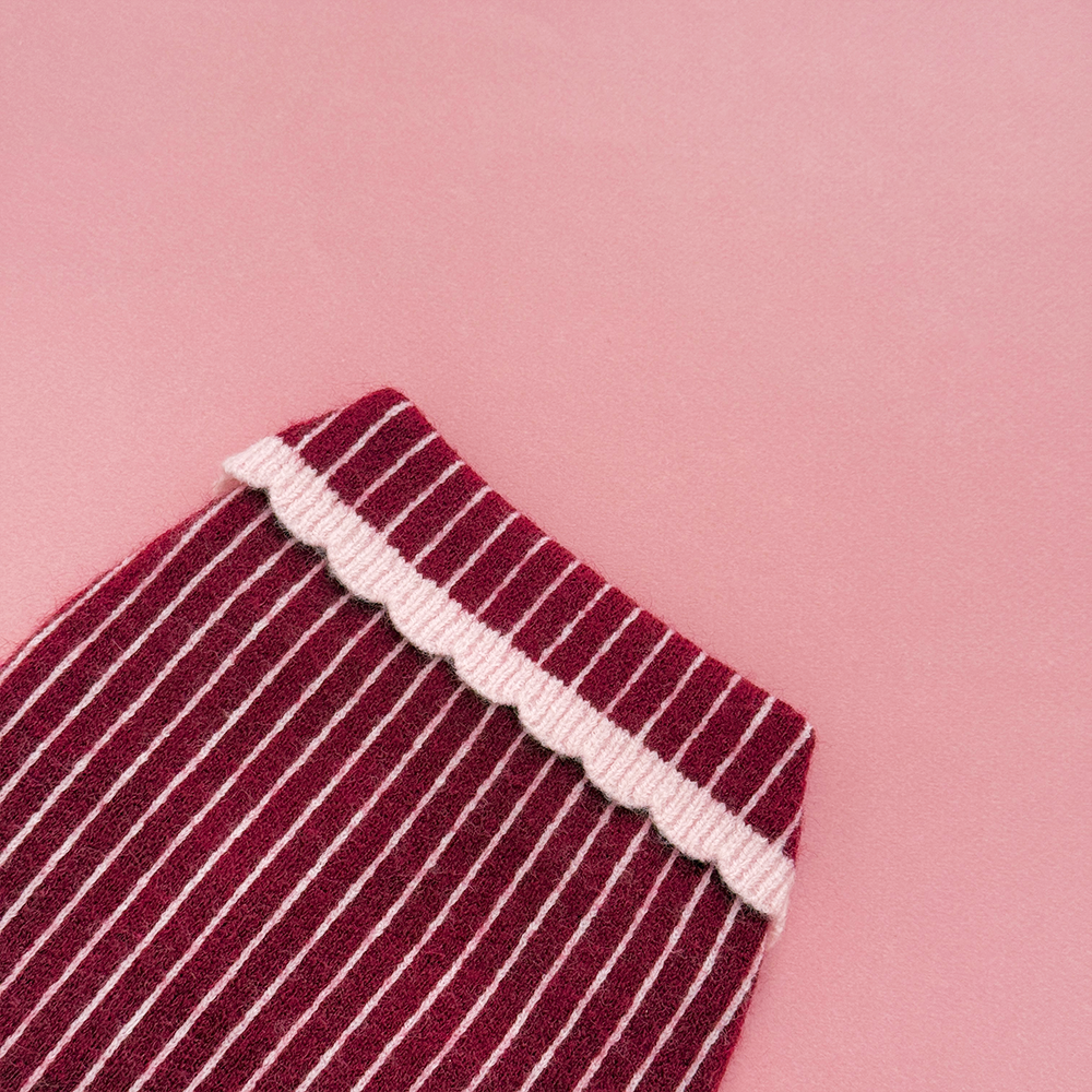 Close up of knitted dog jumper in burgundy with thin baby pink vertical stripes, featuring a pink collar with scalloped edging.