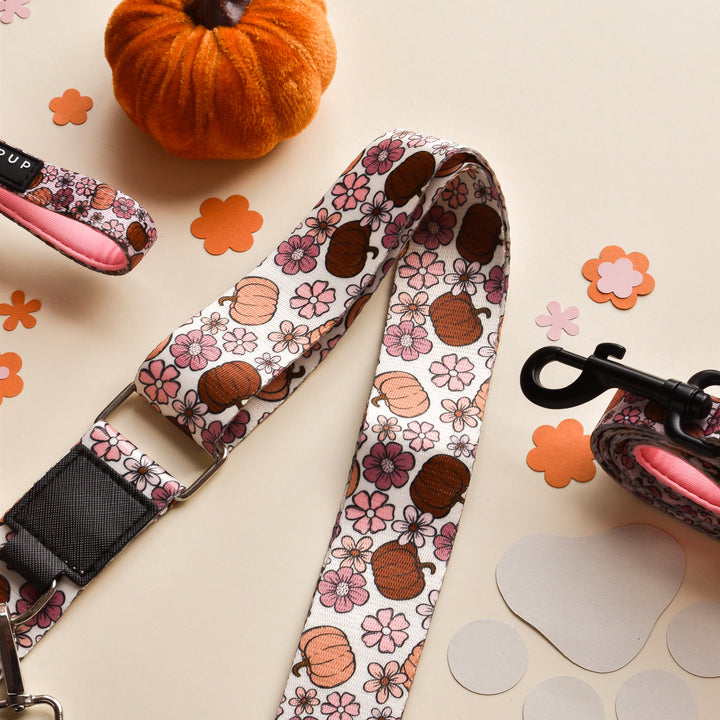 A festive Bag Strap adorned with orange pumpkins and pink flowers is laid out on a light surface. Surrounding the Pup-kin Meadow strap by Cocopup London are flower-shaped confetti and a small plush pumpkin decoration, creating a cheerful autumn theme perfect for Halloween.