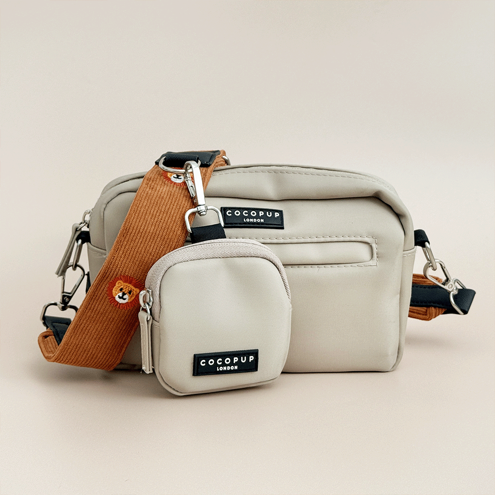 A light beige Cocopup Dog Walking Bag with a matching small treat pouch and a tan colour strap with lion embroidery, photographed on a plain warm beige studio background.