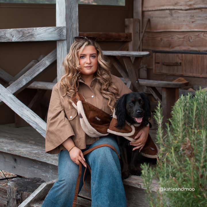 A woman sits on rustic wooden steps holding a black dog wearing a brown and beige shearling-lined jacket. The woman wears a matching brown button-up shirt and blue jeans.