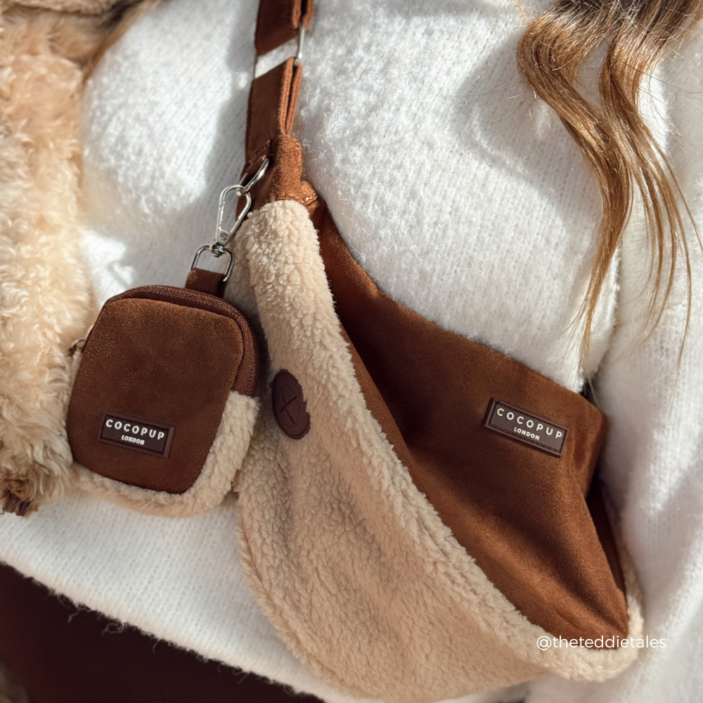 A close-up lifestyle shot of a person wearing a brown suede shoulder bag with a beige sherpa lining and a matching small crossbody pouch. The person is wearing a white knit sweater, and a fluffy cream-colored dog is visible on the left side.
