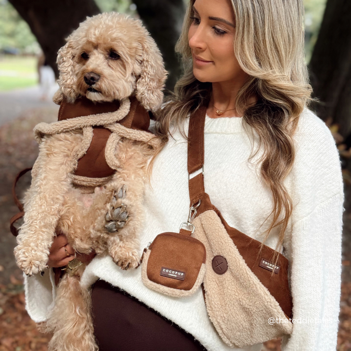 A woman in a white sweater carries a fluffy cream-colored dog wearing a brown harness while wearing a matching brown and cream crossbody dog bag with a small pouch. The brand name 'Cocopup' is visible on the bag's patch and strap label.