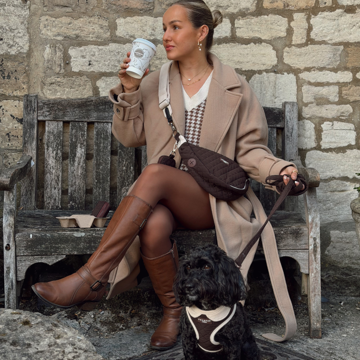 A woman sits on a weathered wooden bench in front of a stone wall, holding a coffee cup and wearing a beige coat, brown boots, and a crossbody bag. A small black dog wearing a harness sits in front of her on a stone surface.