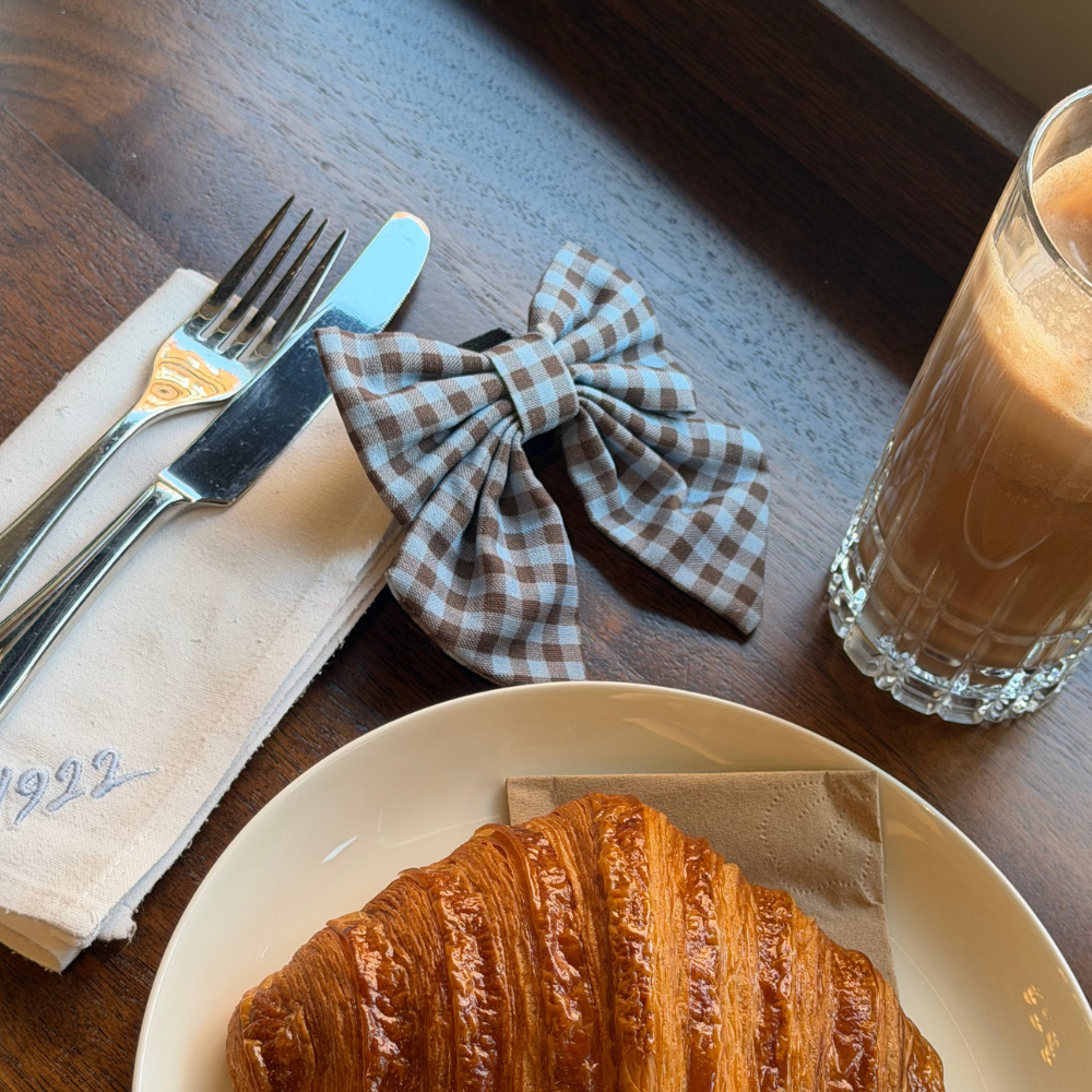 A brown and blue check print sailor bow tie for dogs. With velcro fastening. On a table in a cafe with an iced coffee and croissant