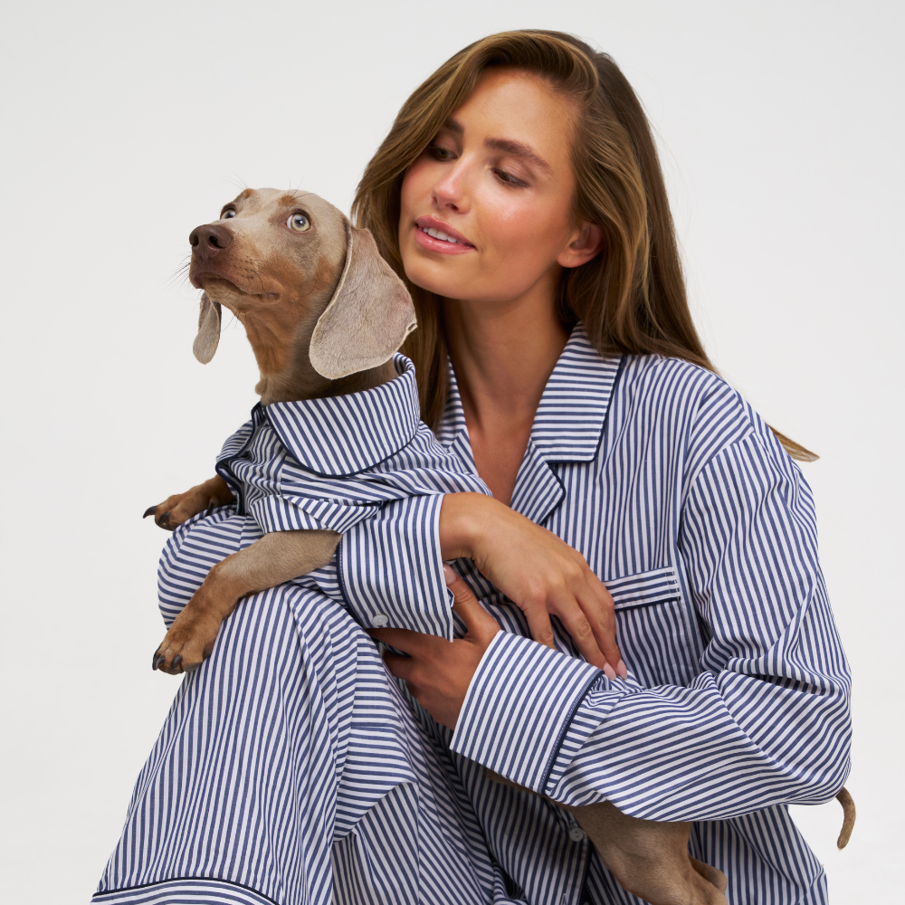 A woman in blue and white striped pajamas holds a small dog wearing a matching pajama shirt against a plain light gray studio background.