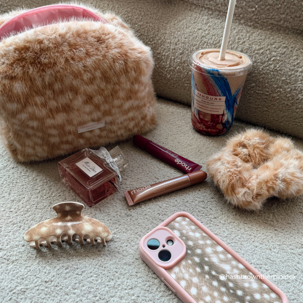 A styled flat-lay of a fluffy faux-fur cosmetic pouch in a warm beige and brown pattern, shown alongside a pink phone case, hair accessories, a perfume bottle, and an iced coffee on a carpeted floor.