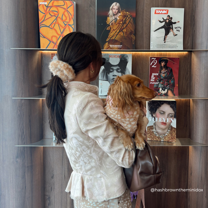 A woman in a cream-colored textured jacket and patterned skirt holds a small golden dachshund dog wearing a pink patterned sweater. They are standing in front of a wooden wall with built-in shelves displaying fashion magazines.