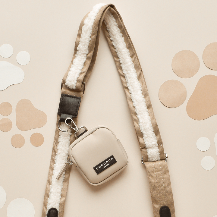 A stylish beige and white crossbody dog walking bag with a sheepskin textured "Nude Aviator" strap from Cocopup London is pictured on a light brown background with paw print and dot designs. The bag includes a small attached pouch featuring the brand label reading "Cocopup London.