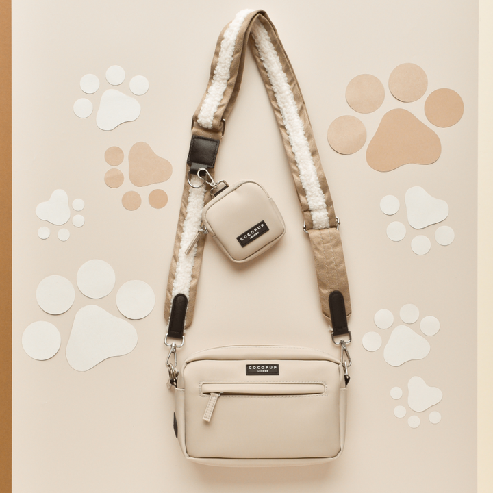 A stylish beige crossbody bag, perfect as a Cocopup Dog Walking Bag, and a matching small pouch are displayed on a tan background adorned with white and brown paw prints. The bag features the "Nude Aviator" faux shearling strap from Cocopup London, and both pieces have black label accents.