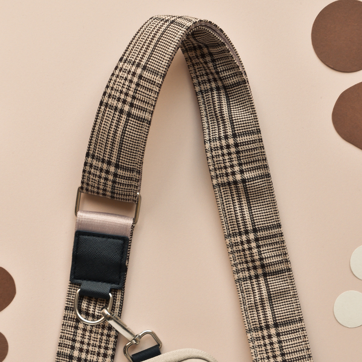 A close-up of the chic Bag Strap - Pup Plaid from Cocopup London attached to a bag. The background is beige with decorative brown and white circles. The strap features a metal clasp and black leather accents, perfect for your Cocopup Dog Walking Bag.