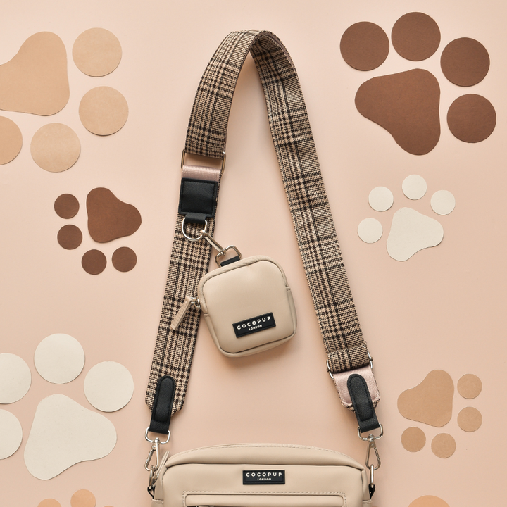 A beige crossbody bag featuring the "Bag Strap - Pup Plaid" from Cocopup London is displayed on a light peach background. A smaller, matching accessory pouch is attached to the strap. The background features various paw print cutouts in shades of brown and cream. Both bags have "COCOPUP" labels.