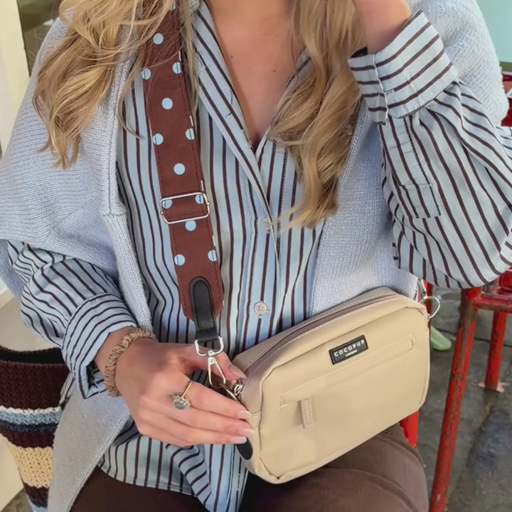 A woman wearing a light blue jumper over a blue and brown striped button-up shirt, holding a beige crossbody bag with a brown polka-dot strap. The bag has a black label that reads 'cocopup london'.