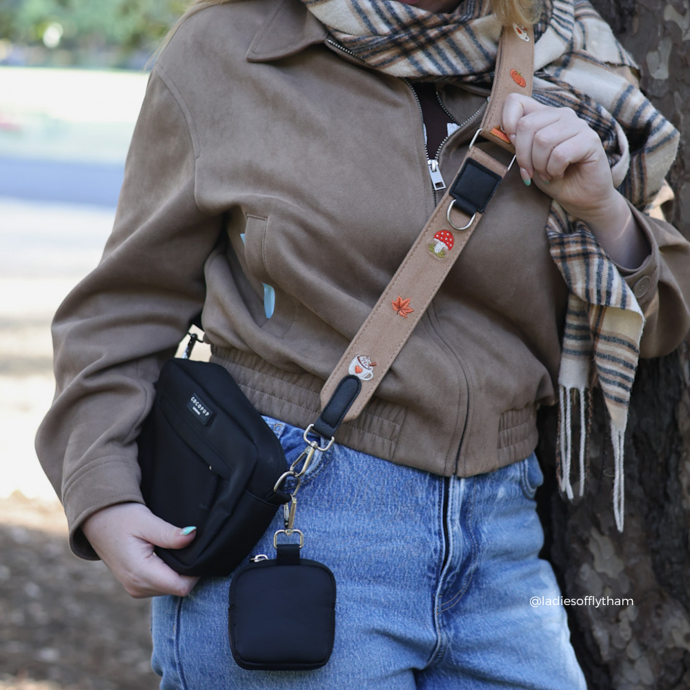 Close-up of a black crossbody bag and treat pouch with a tan suede strap featuring embroidered red and orange mushrooms and a leaf