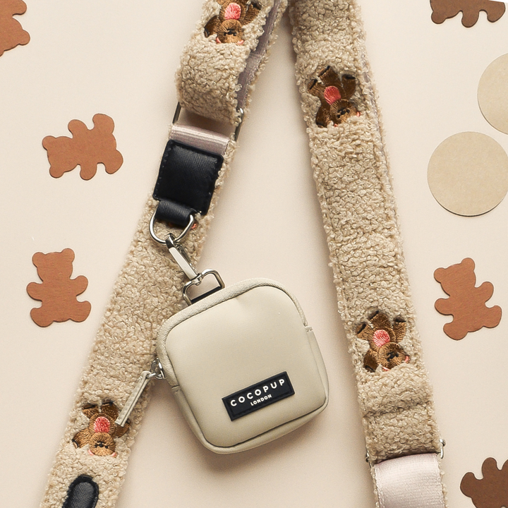 A beige bag strap, named "Theo Embroidered Teddy," with small bear-shaped cutouts, is laid flat on a light background. Attached to the strap is a small beige square pouch labeled "Cocopup London" with a black and white logo, perfect for doubling as a cute dog walking bag.